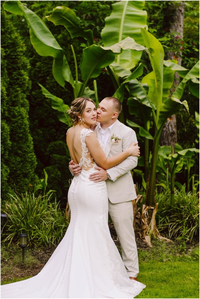 Snohomish Wedding Photographer GSquared Weddings In a lush Snohomish garden, the bride in a white lace gown and groom in a light suit embrace among large green leaves. The bride looks serenely at the camera while the groom gazes lovingly at her, surrounded by vibrant tropical foliage. Serving Seattle, Snohomish and Orlando