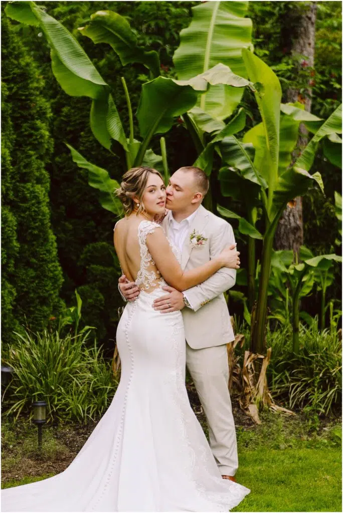 Snohomish Wedding Photographer GSquared Weddings In a lush Snohomish garden, the bride in a white lace gown and groom in a light suit embrace among large green leaves. The bride looks serenely at the camera while the groom gazes lovingly at her, surrounded by vibrant tropical foliage. Serving Seattle, Snohomish and Orlando