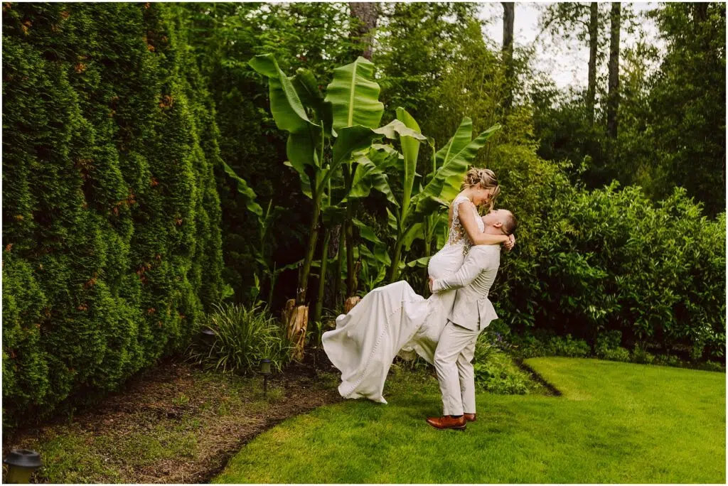 Snohomish Wedding Photographer GSquared Weddings In a lush Snohomish garden, a groom in a light suit lifts the bride in her white dress amid vibrant green foliage and banana plants. Their joy is palpable as she gazes at him lovingly while being held. Serving Seattle, Snohomish and Orlando