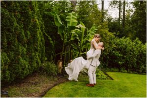 Snohomish Wedding Photographer GSquared Weddings In a lush Snohomish garden, a groom in a light suit lifts the bride in her white dress amid vibrant green foliage and banana plants. Their joy is palpable as she gazes at him lovingly while being held. Serving Seattle, Snohomish and Orlando