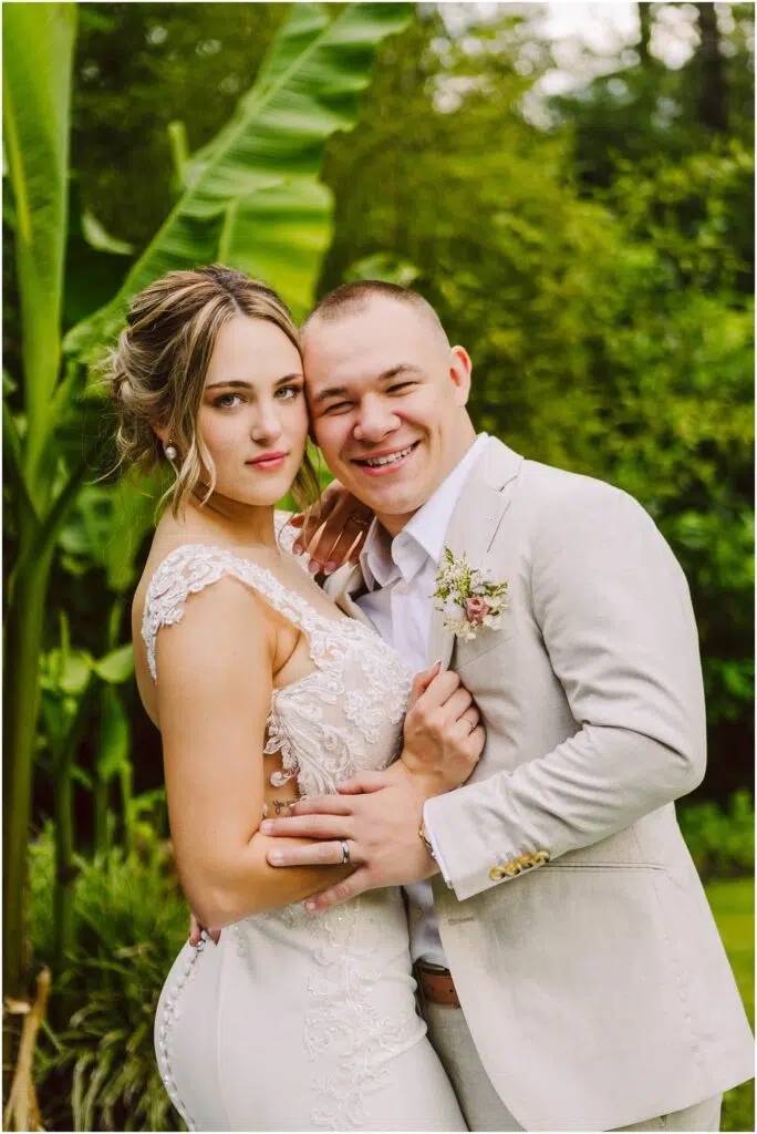 Snohomish Wedding Photographer GSquared Weddings A couple in wedding attire poses outdoors in Snohomish. The woman wears a lace wedding dress, and the man is in a light beige suit with a boutonniere. They are surrounded by lush green foliage, including large banana leaves, and both are smiling. Serving Seattle, Snohomish and Orlando