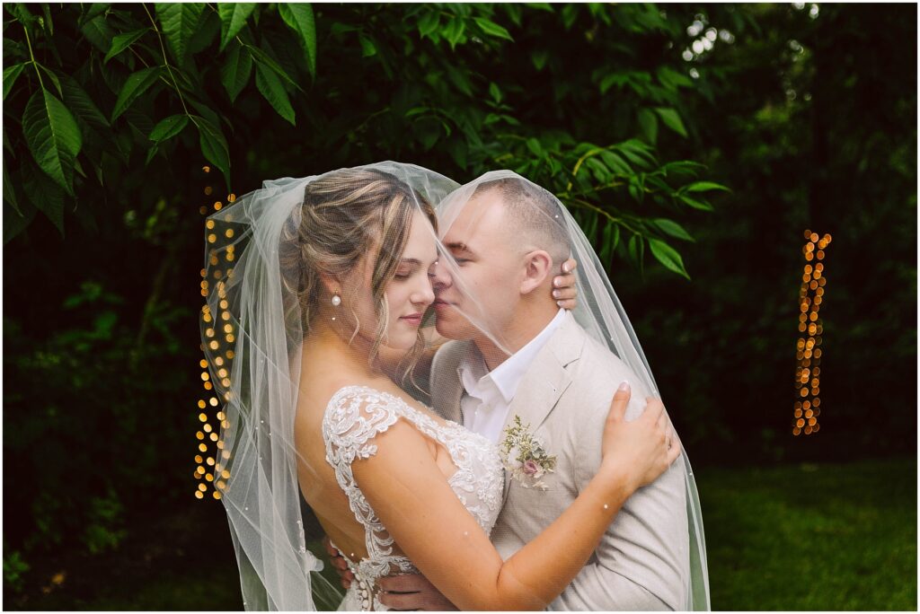 Snohomish Wedding Photographer GSquared Weddings In Snohomish, a bride and groom embrace lovingly under a shared veil. The bride wears a detailed lace wedding dress while the groom dons a light-colored suit. They are enveloped by lush green foliage, with warm, out-of-focus lights twinkling in the background. Serving Seattle, Snohomish and Orlando
