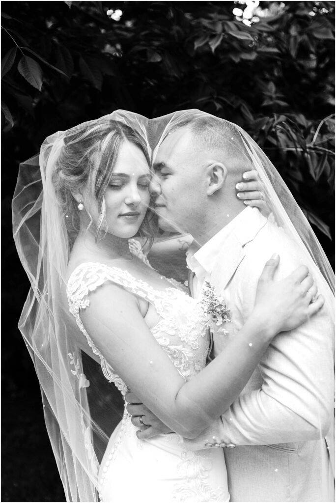 Snohomish Wedding Photographer GSquared Weddings Black and white photo of a couple embracing on their wedding day in Snohomish. The bride is in a lace gown and veil, while the groom sports a light-colored suit. Theyre surrounded by leafy trees, creating a romantic, intimate moment. Serving Seattle, Snohomish and Orlando