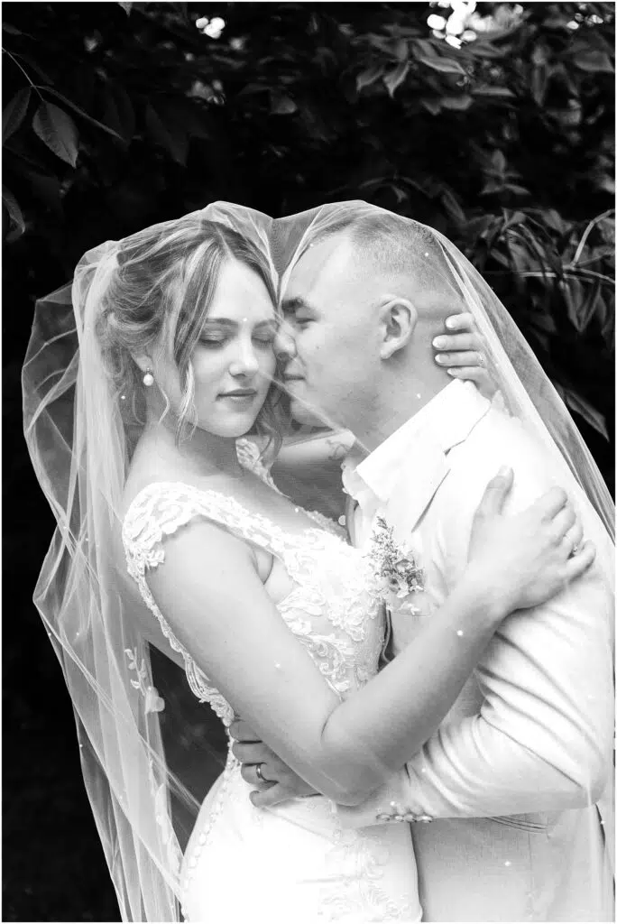 Snohomish Wedding Photographer GSquared Weddings Black and white photo of a couple embracing on their wedding day in Snohomish. The bride is in a lace gown and veil, while the groom sports a light-colored suit. Theyre surrounded by leafy trees, creating a romantic, intimate moment. Serving Seattle, Snohomish and Orlando