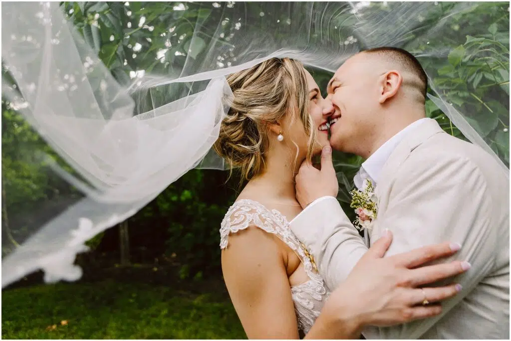 Snohomish Wedding Photographer GSquared Weddings Bride and groom sharing a joyful moment outdoors in Snohomish. The bride’s veil drapes over them as they lean in for a kiss, smiling warmly. The groom holds the bride’s face amid the lush greenery that beautifully defines this picturesque setting. Serving Seattle, Snohomish and Orlando
