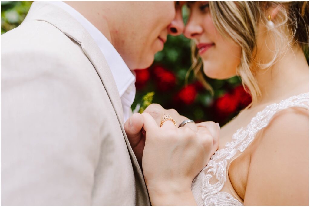 Snohomish Wedding Photographer GSquared Weddings A couple stands close in Snohomish, touching foreheads and holding hands gently. The womans engagement ring is visible. Both are wearing light-colored clothing, with blurred red flowers in the background, creating a romantic atmosphere. Serving Seattle, Snohomish and Orlando