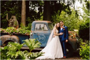 Stanwood Backyard Wedding Snohomish Wedding Photographer GSquared Weddings In a charming Stanwood setting, a bride in a white dress and a groom in a blue suit stand happily in front of a vintage blue pickup truck adorned with flowers. The truck is parked amid the lush beauty of their backyard garden, creating the perfect backdrop for their wedding day. Serving Seattle, Snohomish and Orlando