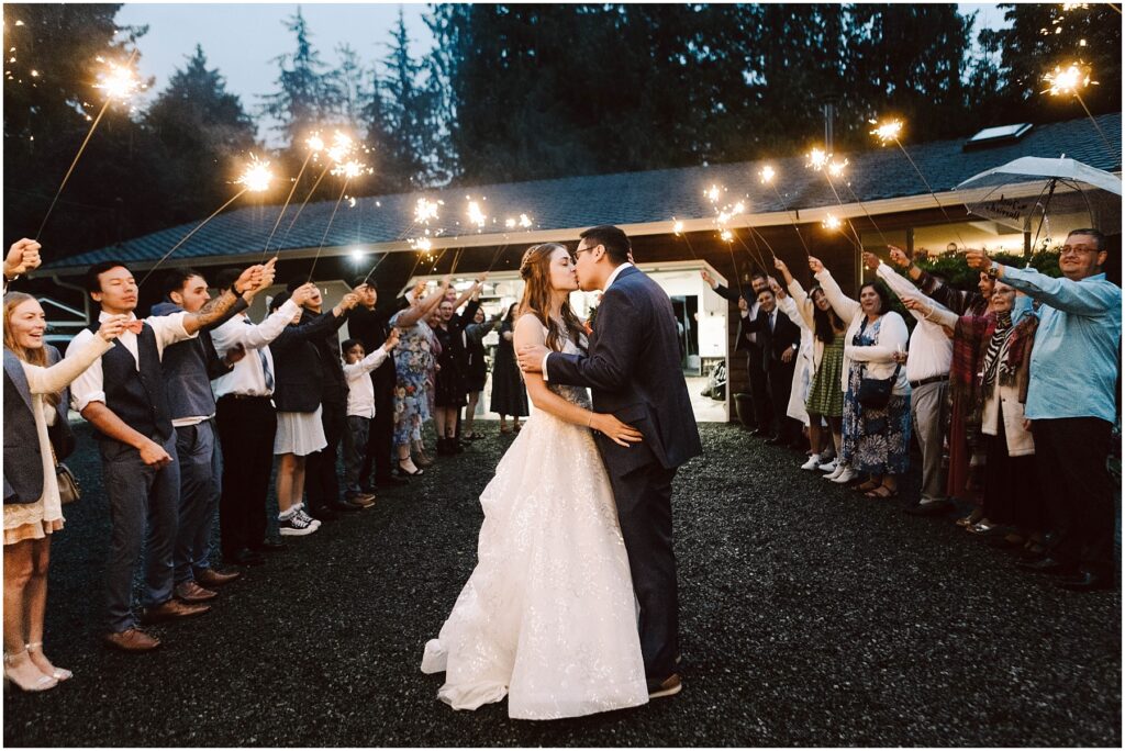 Snohomish Wedding Photographer GSquared Weddings A bride and groom share a kiss under the night sky during their backyard wedding, surrounded by guests holding sparklers. The scene is festive, with an arch of light formed by the guests and a backdrop of trees and a lit building. Serving Seattle, Snohomish and Orlando