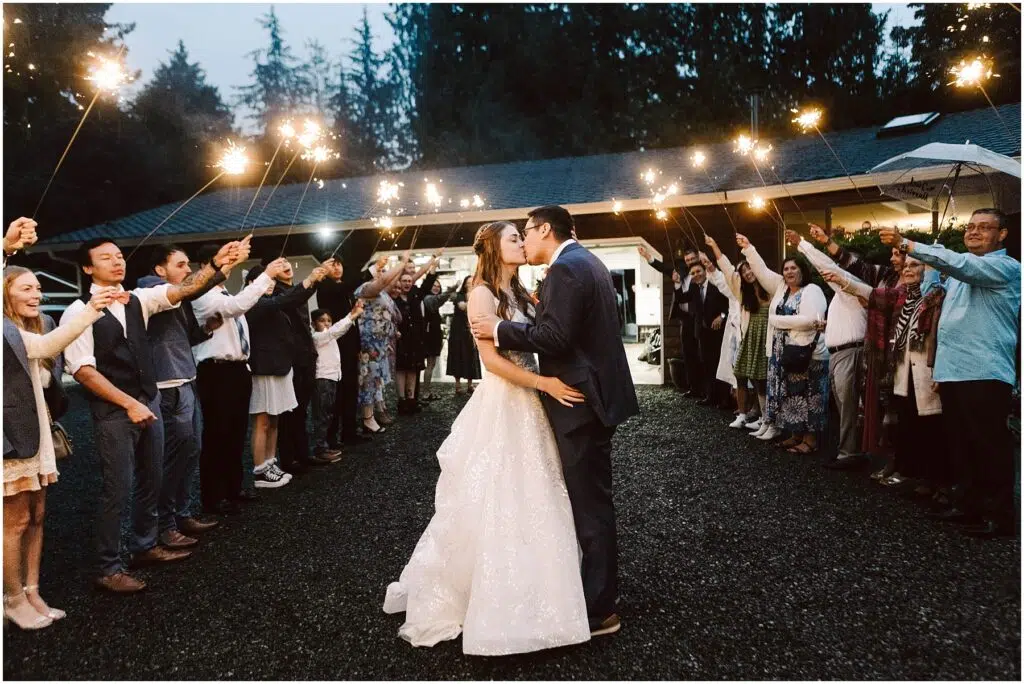 Snohomish Wedding Photographer GSquared Weddings A bride and groom share a kiss under the night sky during their backyard wedding, surrounded by guests holding sparklers. The scene is festive, with an arch of light formed by the guests and a backdrop of trees and a lit building. Serving Seattle, Snohomish and Orlando