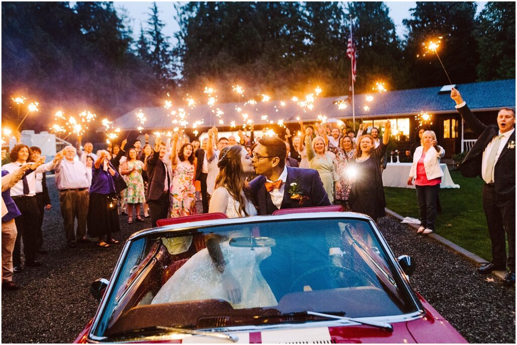 Snohomish Wedding Photographer GSquared Weddings A bride and groom kiss while sitting in a red convertible during a magical nighttime backyard wedding celebration. Guests stand around holding sparklers, with a lit building visible in the background amidst the trees. Serving Seattle, Snohomish and Orlando