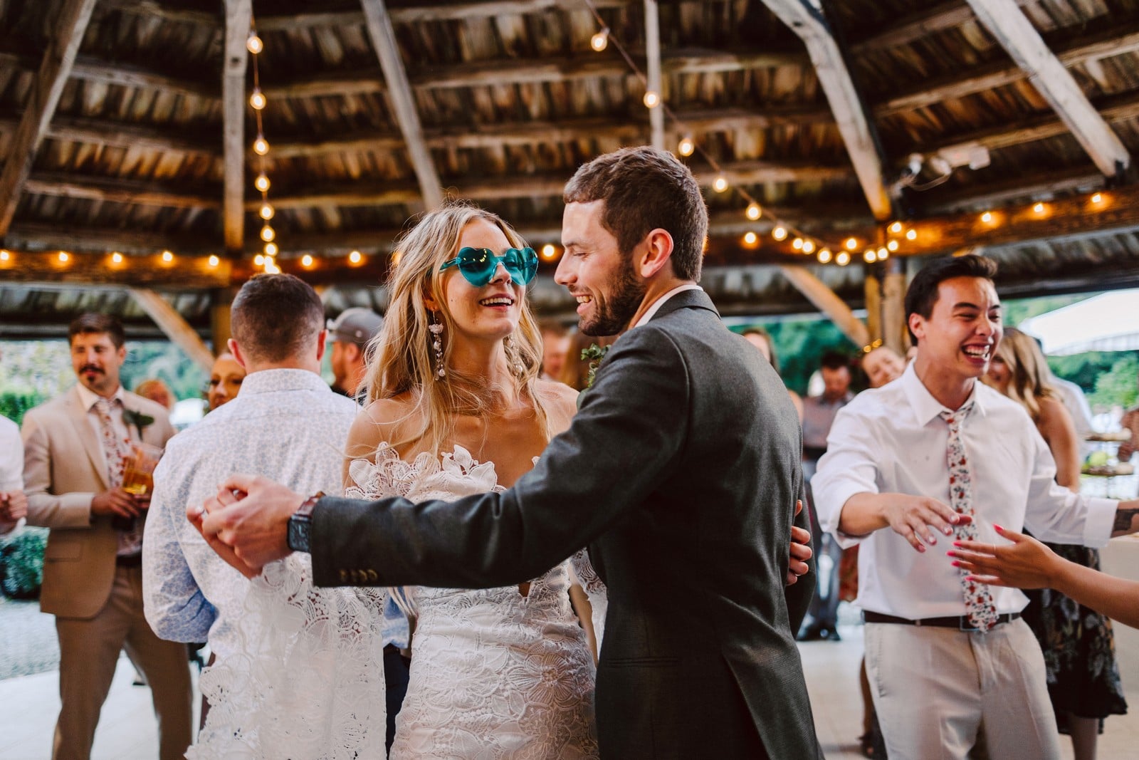 GSquared Weddings Photography A bride in a white dress and blue sunglasses dances with a man in a suit under string lights, surrounded by smiling guests at their outdoor Snohomish wedding celebration—captured by expert weddings photography. GSquared Weddings | 47.9129° N, 122.0982° W | Serving Snohomish, Seattle , Orlando & Beyond