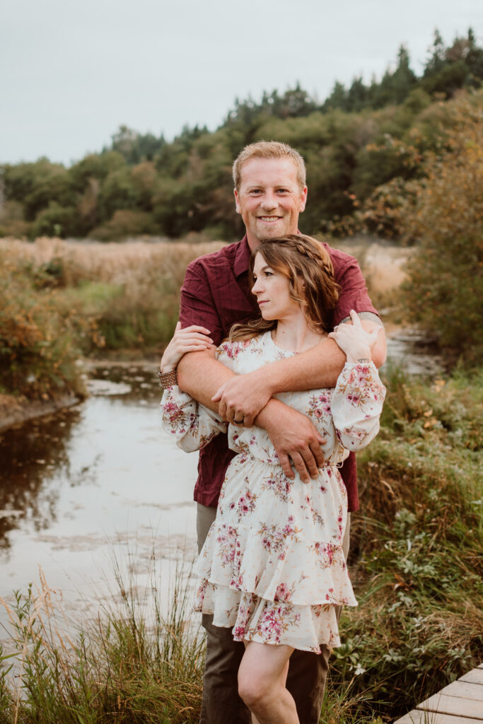 GSquared Weddings Photography A man in a maroon shirt hugs a woman in a white floral dress from behind. By a stream with tall grass and trees, they look content and peaceful—perfect for capturing on your Elementor website to highlight serene, romantic moments. GSquared Weddings | 47.9129° N, 122.0982° W | Serving Snohomish, Seattle , Orlando & Beyond