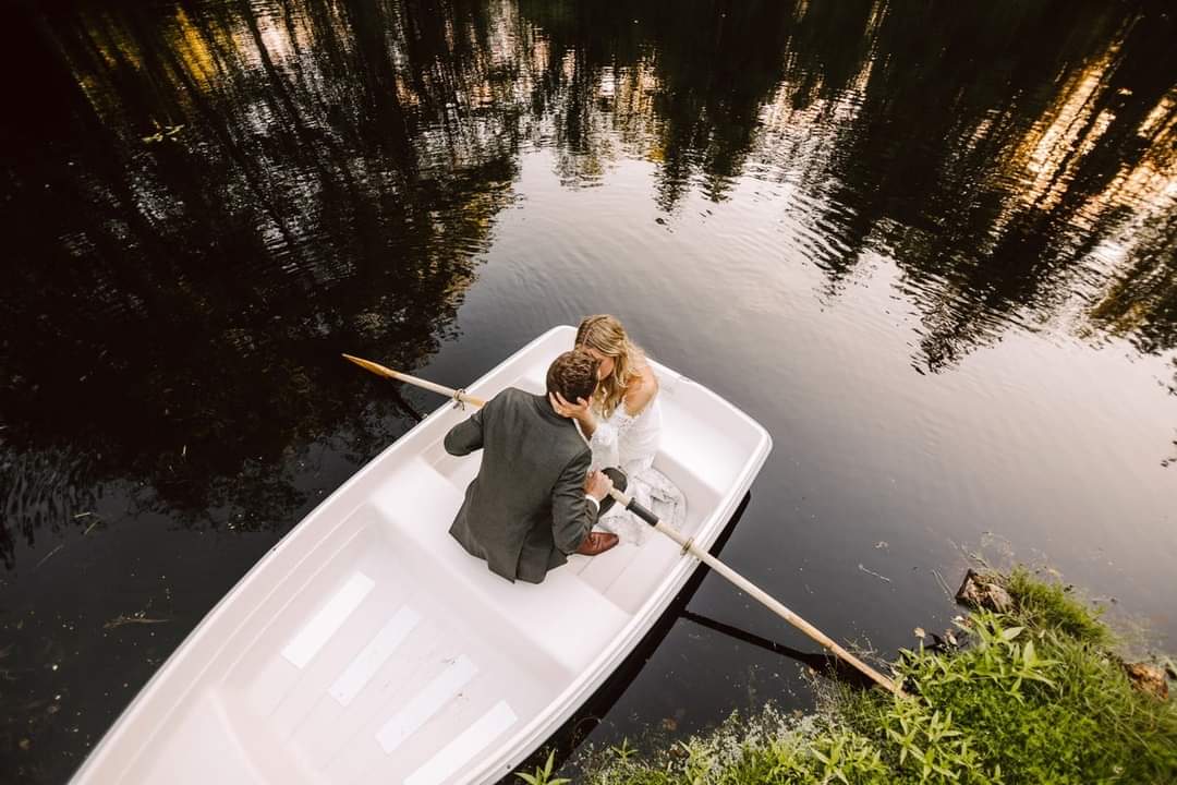 GSquared Weddings Photography A couple dressed in wedding attire sits closely together in a white rowboat on a calm, reflective lake, surrounded by greenery and trees, with the groom gently holding the bride—captured beautifully by GSquared Weddings Photography. GSquared Weddings | 47.9129° N, 122.0982° W | Serving Snohomish, Seattle , Orlando & Beyond