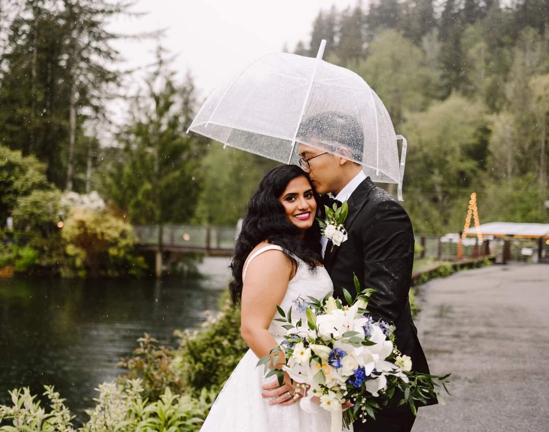GSquared Weddings Photography A bride and groom stand close together under a clear umbrella by a lake near Snohomish, surrounded by greenery. The groom kisses the bride’s forehead as she smiles, holding a white and blue bouquet—a perfect moment of Seattle weddings photography on a rainy day. GSquared Weddings | 47.9129° N, 122.0982° W | Serving Snohomish, Seattle , Orlando & Beyond