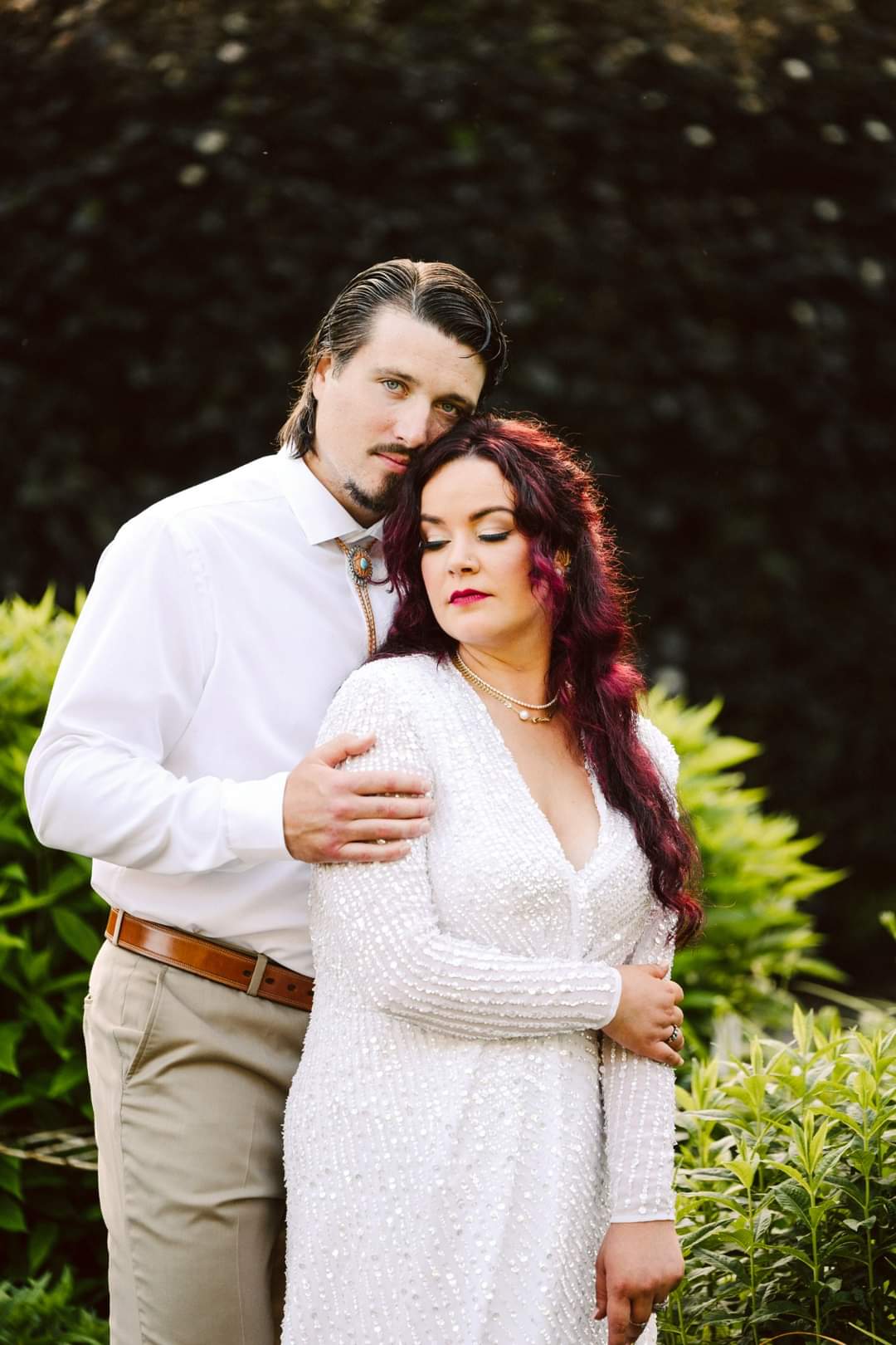 GSquared Weddings Photography A man in a white shirt and khaki pants lovingly embraces a woman in a beaded dress with long, wavy hair. Surrounded by green bushes, they capture a tender moment—an example of heartfelt Snohomish weddings photography. GSquared Weddings | 47.9129° N, 122.0982° W | Serving Snohomish, Seattle , Orlando & Beyond