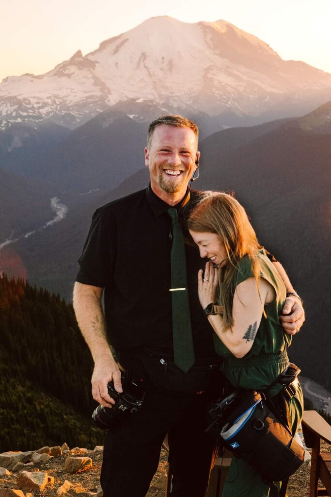 GSquared Weddings Photography A man and woman stand close together, smiling and laughing, on a mountain overlook at sunset. Snow-capped peaks and forested hills create a scenic background—perfect inspiration for an About page designed in WordPress with Elementor. Both wear camera gear, suggesting they are photographers. GSquared Weddings | 47.9129° N, 122.0982° W | Serving Snohomish, Seattle , Orlando & Beyond
