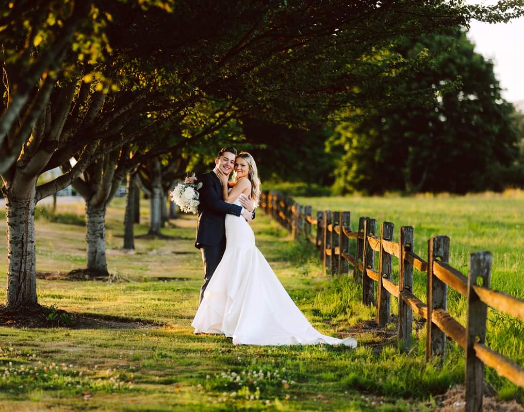 GSquared Weddings Photography A bride and groom embrace and smile under a row of trees by a wooden fence in a sunlit outdoor setting, captured beautifully by GSquared Weddings—a perfect moment of wedding photography. GSquared Weddings | 47.9129° N, 122.0982° W | Serving Snohomish, Seattle , Orlando & Beyond