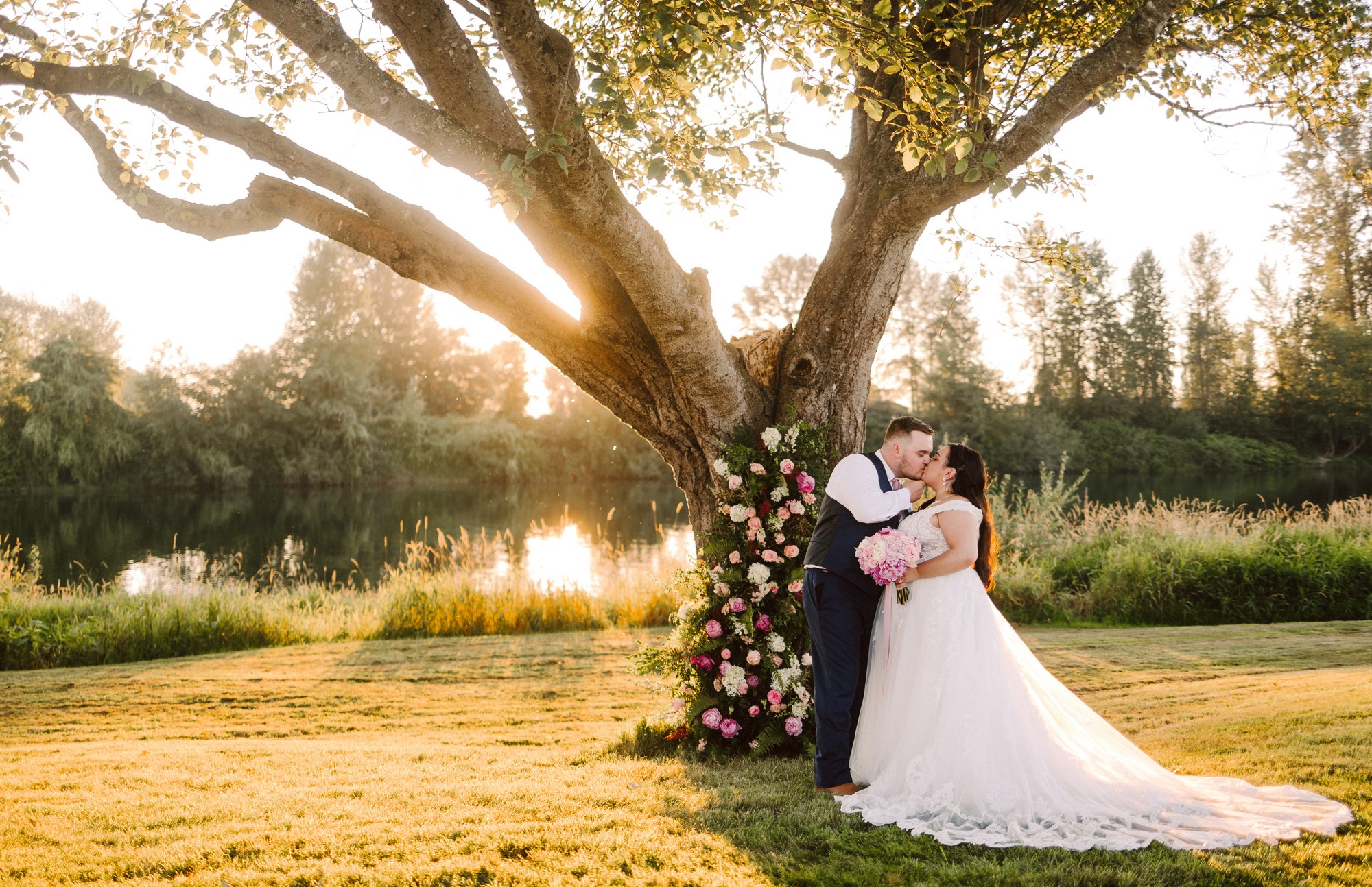 GSquared Weddings Photography A bride and groom kiss under a large tree decorated with flowers by a river at sunset. Captured by GSquared Weddings, the sunlight filters through the branches, creating a romantic, golden wedding photography scene. GSquared Weddings | 47.9129° N, 122.0982° W | Serving Snohomish, Seattle , Orlando & Beyond