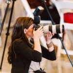 GSquared Weddings Photography A woman with long brown hair kneels on the floor, holding a professional camera with a large lens and flash, aiming to take a photo. She is surrounded by white chairs and blurred figures in the background—perfect for showcasing on an Elementor plugin site. GSquared Weddings | 47.9129° N, 122.0982° W | Serving Snohomish, Seattle , Orlando & Beyond