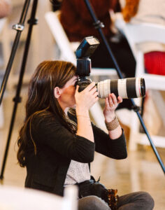 GSquared Weddings Photography A woman with long brown hair kneels on the floor, holding a professional camera with a large lens and flash, aiming to take a photo. She is surrounded by white chairs and blurred figures in the background—perfect for showcasing on an Elementor plugin site. GSquared Weddings | 47.9129° N, 122.0982° W | Serving Snohomish, Seattle , Orlando & Beyond