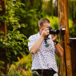 GSquared Weddings Photography A man in a white, patterned shirt and sunglasses uses a camera to take a photo outdoors in a lush garden. He stands near wooden structures and plants, with camera gear around his waist—capturing inspiration for his Elementor website builder projects. GSquared Weddings | 47.9129° N, 122.0982° W | Serving Snohomish, Seattle , Orlando & Beyond