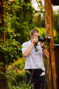 GSquared Weddings Photography A man in a white, patterned shirt and sunglasses uses a camera to take a photo outdoors in a lush garden. He stands near wooden structures and plants, with camera gear around his waist—capturing inspiration for his Elementor website builder projects. GSquared Weddings | 47.9129° N, 122.0982° W | Serving Snohomish, Seattle , Orlando & Beyond