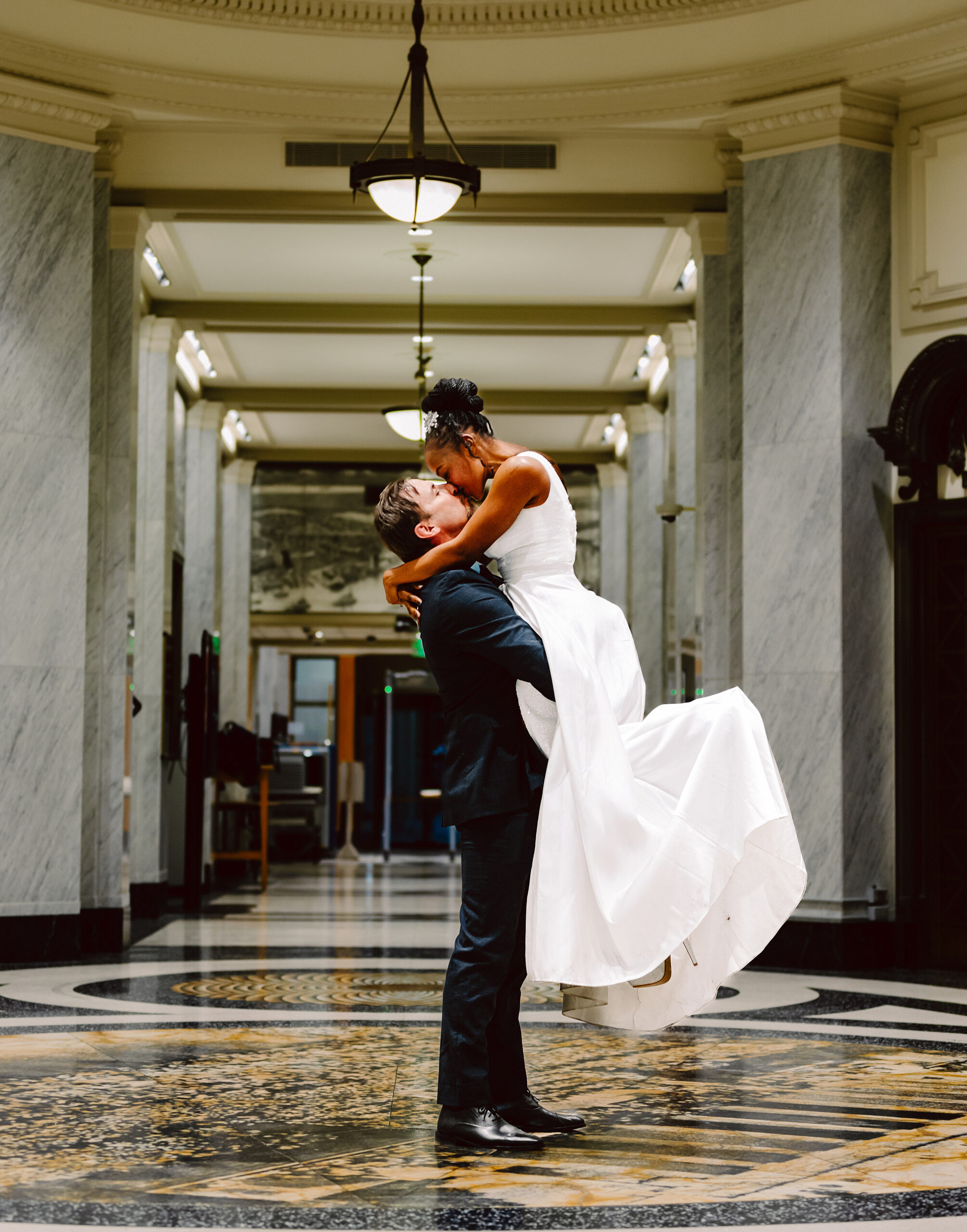Seattle Courthouse Elopement Couple in the Hallway Seattle Courthouse Elopement GSquared Weddings Photography A groom in a dark suit lifts and embraces his bride who wears a white dress in an elegant hallway with marble columns and ornate floorscapturing the joy and intimacy often found at celebrations inspired by affordable elopement prices