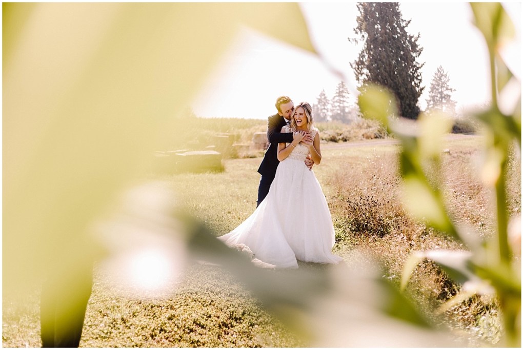 GSquared Weddings Photography A bride and groom stand together outdoors, smiling and embracing in a sunlit field, framed by blurred green leaves in the foreground—a joyful moment captured by GSquared Weddings with trees softly glowing behind. GSquared Weddings | 47.9129° N, 122.0982° W | Serving Snohomish, Seattle , Orlando & Beyond