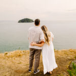 GSquared Weddings Photography A couple stands arm in arm on a grassy cliff overlooking the ocean, facing a small island in the distance. The woman wears a flowing white dress and the man wears a striped shirt and pants. 47.9129° N, 122.0982° W | Serving Snohomish, Seattle , Orlando & Beyond