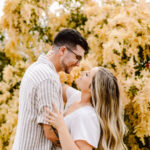 GSquared Weddings Photography A couple smiles lovingly at each other in front of a bush covered in yellow flowers. The woman, in a white dress, wraps her arms around the man, who wears glasses and a striped shirt. 47.9129° N, 122.0982° W | Serving Snohomish, Seattle , Orlando & Beyond