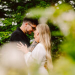 GSquared Weddings Photography A couple stands close together outdoors, touching foreheads and smiling softly, surrounded by greenery and blurred foliage in the foreground. 47.9129° N, 122.0982° W | Serving Snohomish, Seattle , Orlando & Beyond