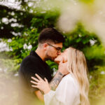 GSquared Weddings Photography A couple stands outdoors surrounded by greenery, sharing a kiss. The woman has long blonde hair and wears a white dress, while the man has dark hair, glasses, and a tattooed arm, wearing a black shirt. Blurred foliage is in the foreground. 47.9129° N, 122.0982° W | Serving Snohomish, Seattle , Orlando & Beyond