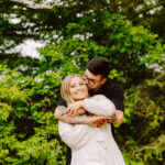 GSquared Weddings Photography A smiling woman in a white dress stands in front of green trees as a man in a black shirt hugs her from behind and kisses her cheek. They both look happy and relaxed outdoors. 47.9129° N, 122.0982° W | Serving Snohomish, Seattle , Orlando & Beyond