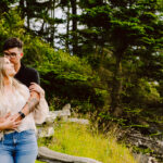 GSquared Weddings Photography A couple stands close together by a rustic wooden fence, surrounded by greenery and trees near a rocky shoreline. The man hugs the woman from behind, both looking content and relaxed outdoors. 47.9129° N, 122.0982° W | Serving Snohomish, Seattle , Orlando & Beyond