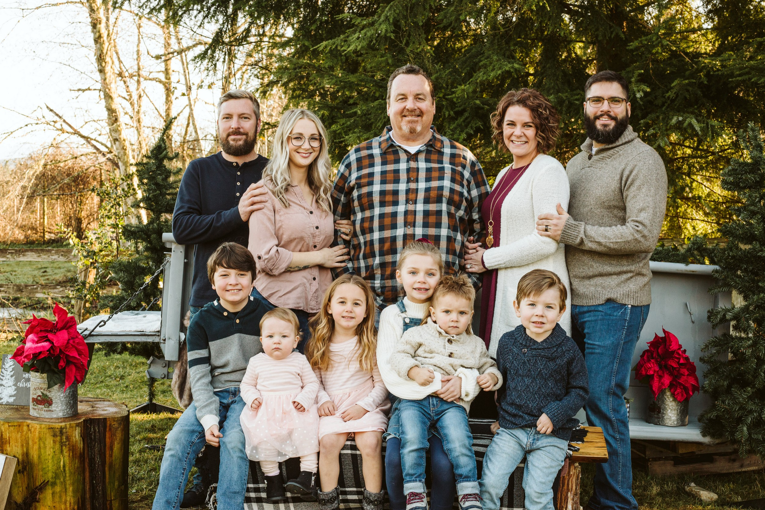 GSquared Weddings Photography A group of four adults and six children pose outdoors for a family photo in front of evergreens. The adults stand behind a bench where the children sit. Everyone is smiling, and there are two red poinsettias on either side. 47.9129° N, 122.0982° W | Serving Snohomish, Seattle , Orlando & Beyond