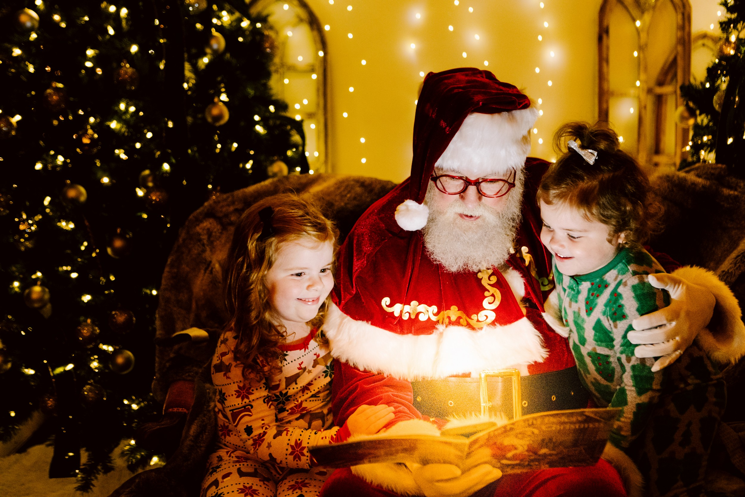 GSquared Weddings Photography Santa Claus sits on a cozy chair between two smiling children, reading a book together. Warm Christmas lights and a decorated tree create a festive, magical atmosphere in the background. 47.9129° N, 122.0982° W | Serving Snohomish, Seattle , Orlando & Beyond