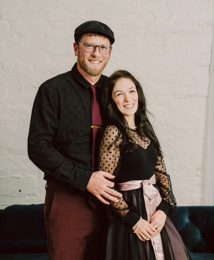 Snohomish Wedding Photographer GSquared Weddings A smiling couple poses together indoors. The man wears glasses, a flat cap, a black shirt, and maroon pants. The woman, about his height, wears a long black dress with sheer polka-dot sleeves and a pink sash. They stand in front of a white brick wall. Serving Seattle, Snohomish and Orlando