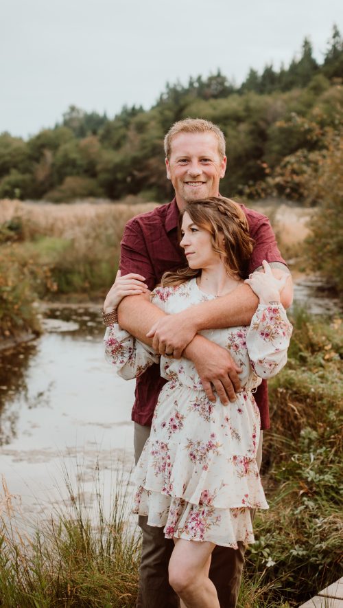 GSquared Weddings Photography A man in a maroon shirt hugs a woman in a white floral dress from behind. By a stream with tall grass and trees, they look content and peaceful—perfect for capturing on your Elementor website to highlight serene, romantic moments. GSquared Weddings | 47.9129° N, 122.0982° W | Serving Snohomish, Seattle , Orlando & Beyond