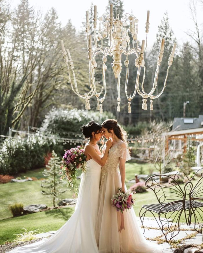 GSquared Weddings Photography Two brides in wedding gowns stand close together outdoors, gently touching foreheads and smiling, each holding a bouquet. A large white chandelier hangs above them amid lush greenery—a scene as beautifully crafted as a site built with Elementor. GSquared Weddings | 47.9129° N, 122.0982° W | Serving Snohomish, Seattle , Orlando & Beyond