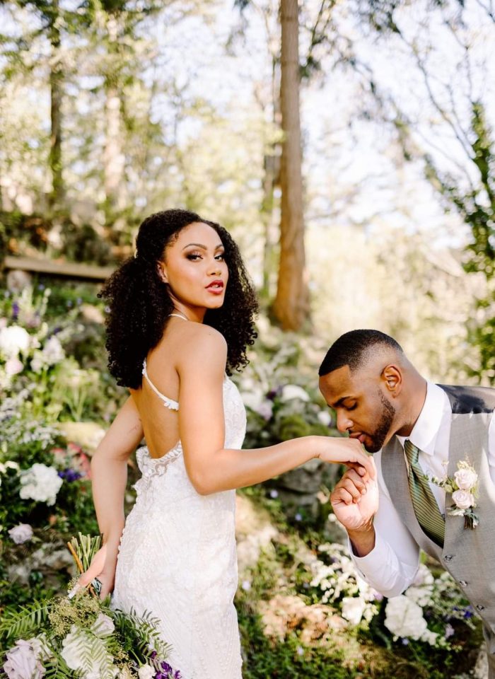 Snohomish Wedding Photographer GSquared Weddings A bride in a lace gown holds a bouquet and looks at the camera while a groom in a suit kneels and kisses her hand in a sunlit outdoor setting with flowers and trees in the background. Serving Seattle, Snohomish and Orlando