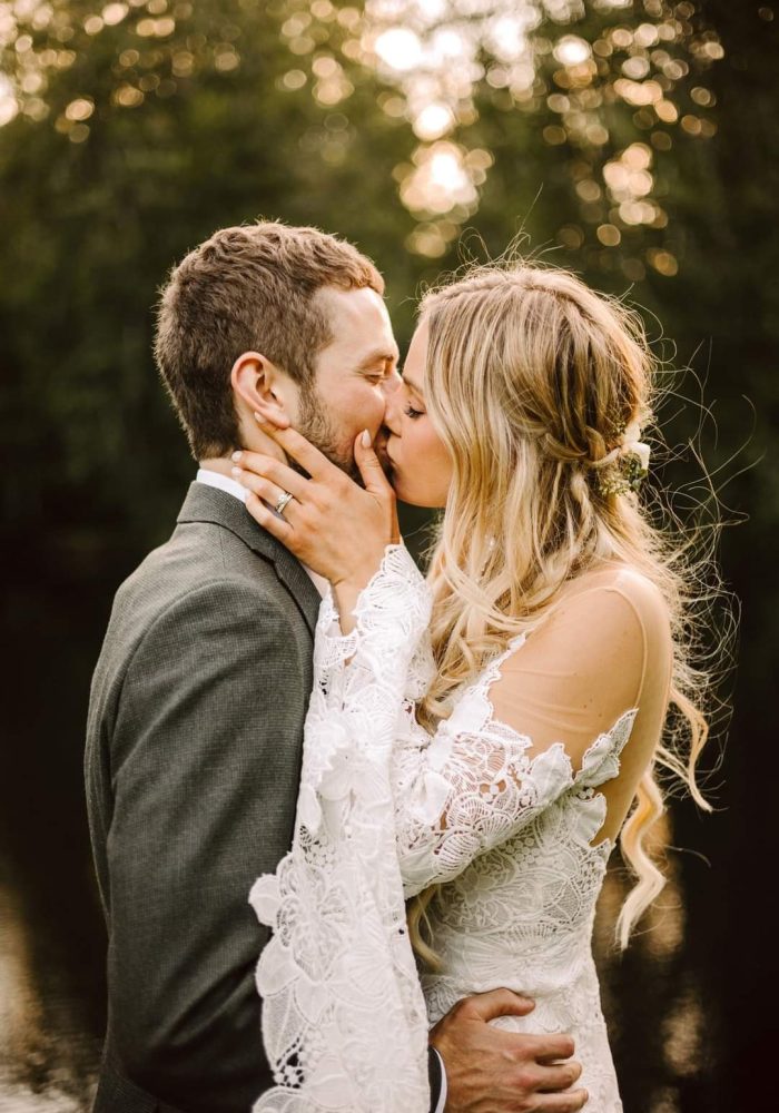 GSquared Weddings Photography A bride and groom share a kiss outdoors in wedding attire during their Full Weddings celebration. The bride wears a lace, long-sleeve dress and her hair in loose waves, while the groom is in a suit beneath trees and soft sunlight. GSquared Weddings | 47.9129° N, 122.0982° W | Serving Snohomish, Seattle , Orlando & Beyond
