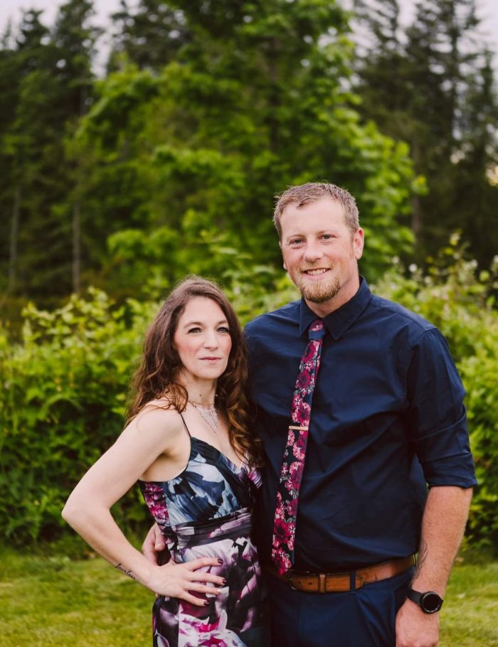 Snohomish Wedding Photographer GSquared Weddings A woman in a floral dress stands next to a man in a navy shirt and floral tie. They smile at the camera, posing outdoors with lush green trees—a beautiful moment captured by GSquared Weddings, specialists in Snohomish wedding photography. Serving Seattle, Snohomish and Orlando