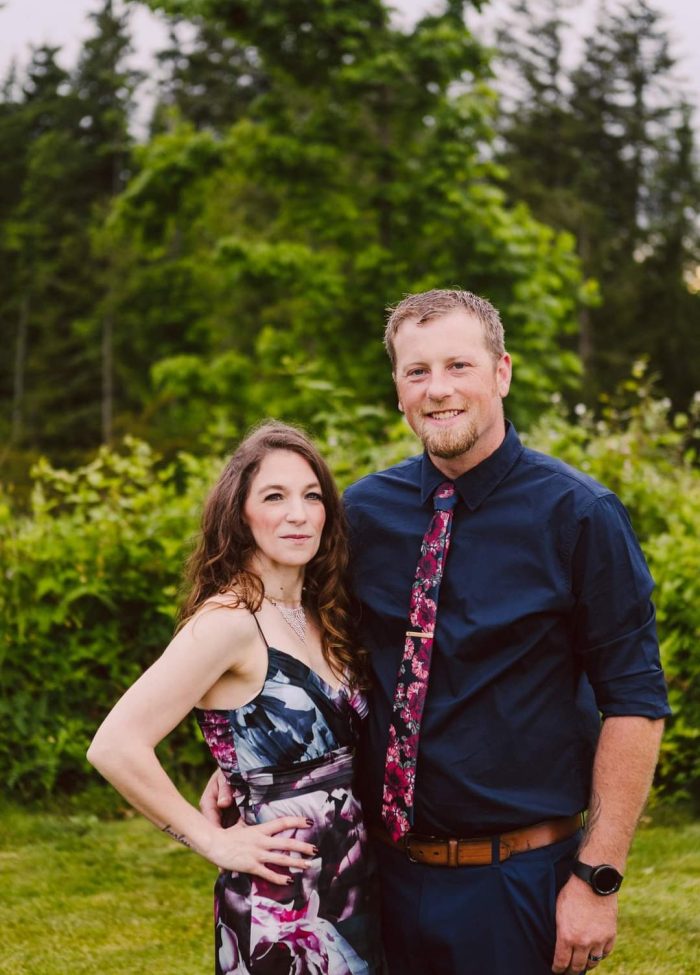 Snohomish Wedding Photographer GSquared Weddings A woman in a floral dress stands next to a man in a navy shirt and floral tie. They smile at the camera, posing outdoors with lush green trees—a beautiful moment captured by GSquared Weddings, specialists in Snohomish wedding photography. Serving Seattle, Snohomish and Orlando