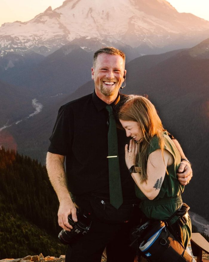 GSquared Weddings Photography A man and woman stand close together, smiling and laughing, on a mountain overlook at sunset. Snow-capped peaks and forested hills create a scenic background—perfect inspiration for an About page designed in WordPress with Elementor. Both wear camera gear, suggesting they are photographers. GSquared Weddings | 47.9129° N, 122.0982° W | Serving Snohomish, Seattle , Orlando & Beyond