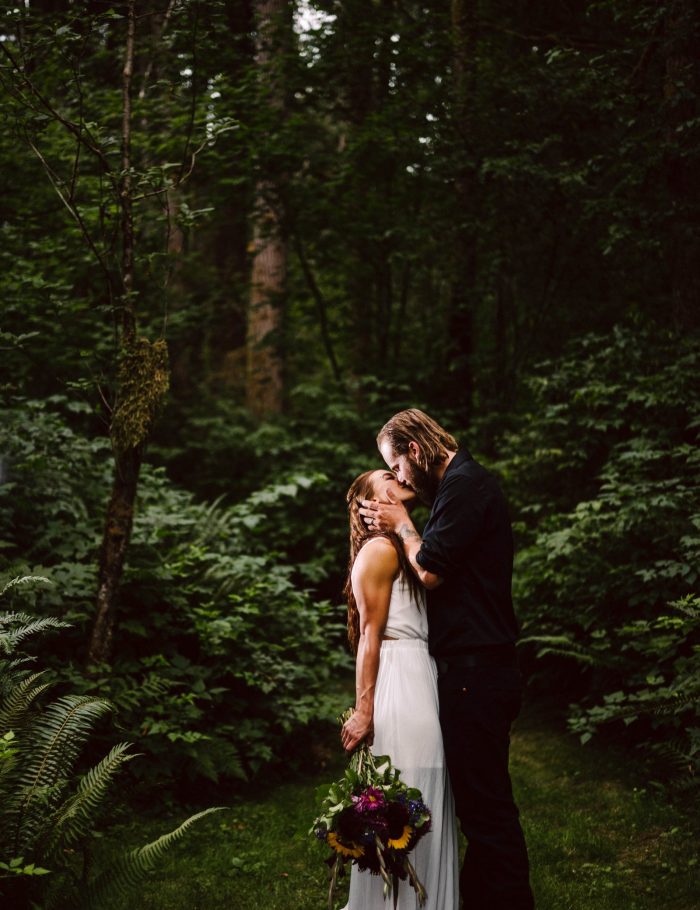 GSquared Weddings Photography A couple stands in a lush, green forest; the woman in a white dress holds a bouquet while the man, dressed in black, gently kisses her forehead. Sunlight filters softly through the trees—an intimate moment that makes elopement packages truly unforgettable. GSquared Weddings | 47.9129° N, 122.0982° W | Serving Snohomish, Seattle , Orlando & Beyond