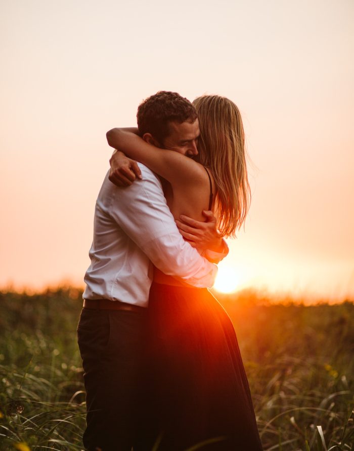 Snohomish Wedding Photographer GSquared Weddings A couple embraces in a grassy field at sunset, with warm sunlight glowing behind them. The woman wears a sleeveless dress, and the man wears a white shirt. They appear affectionate and peaceful in the natural setting. Serving Seattle, Snohomish and Orlando