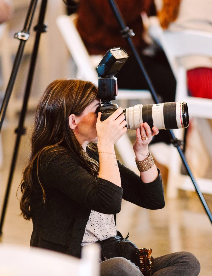 GSquared Weddings Photography A woman with long brown hair kneels on the floor, holding a professional camera with a large lens and flash, aiming to take a photo. She is surrounded by white chairs and blurred figures in the background—perfect for showcasing on an Elementor plugin site. GSquared Weddings | 47.9129° N, 122.0982° W | Serving Snohomish, Seattle , Orlando & Beyond