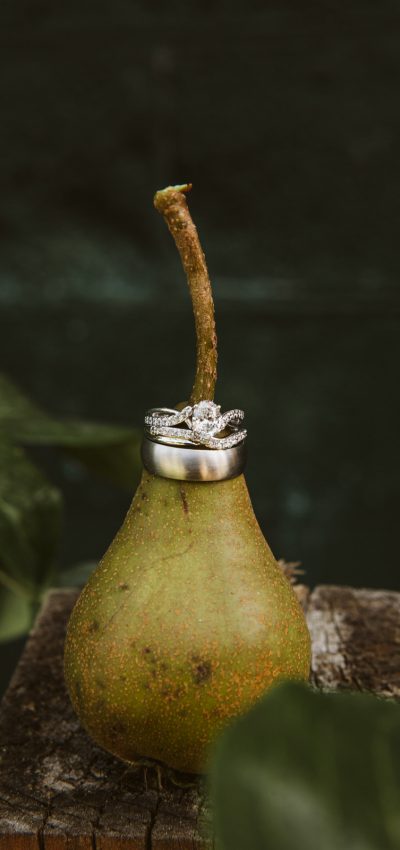 GSquared Weddings Photography A green pear stands upright on a wooden surface with two rings, one with diamonds, stacked around its stem—perfect inspiration for couples exploring elopement prices. The dark, blurred background features hints of green leaves. GSquared Weddings | 47.9129° N, 122.0982° W | Serving Snohomish, Seattle , Orlando & Beyond
