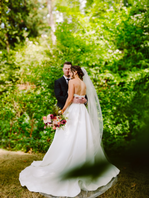 GSquared Weddings Photography A bride in a white gown and veil holds a bouquet while facing a groom in a dark suit. They stand close together outdoors, surrounded by lush green foliage. 47.9129° N, 122.0982° W | Serving Snohomish, Seattle , Orlando & Beyond
