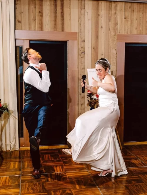 GSquared Weddings Photography A bride in a white dress and a groom in a vest and tie enthusiastically dance together on the wooden floor at their wedding, each lifting one leg high in the air. The bride holds a microphone and smiles. 47.9129° N, 122.0982° W | Serving Snohomish, Seattle , Orlando & Beyond